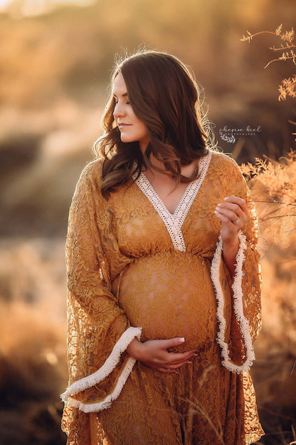 Pregnant model poses outside with a cognac dress made of lace with details in white color on the sleeves and top. The top has a low v cut neckline and kaftan sleeves. She is looking to the side and holds her bump with one hand. 