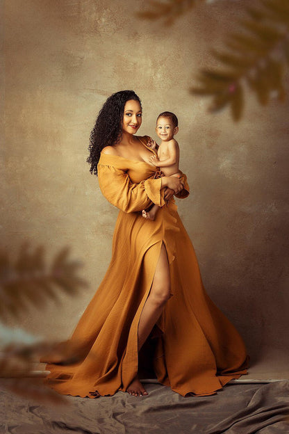 Model poses together with her son during a studio photoshoot. She stares at the front and wears a long dress in ocre color. The dress has a low-v cut neckline and a split on the legs. The split has ruffle details. 
