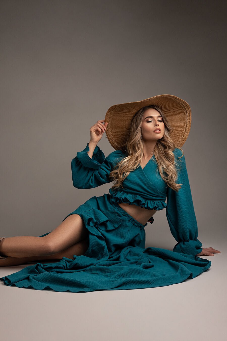 blond model poses laying on the studio floor, leaning with one hand on the floor and the other touching the ending of a big brown hat on her head. she is wearing a boho western chic outfit in petrol color, with long sleeves and ruffle details