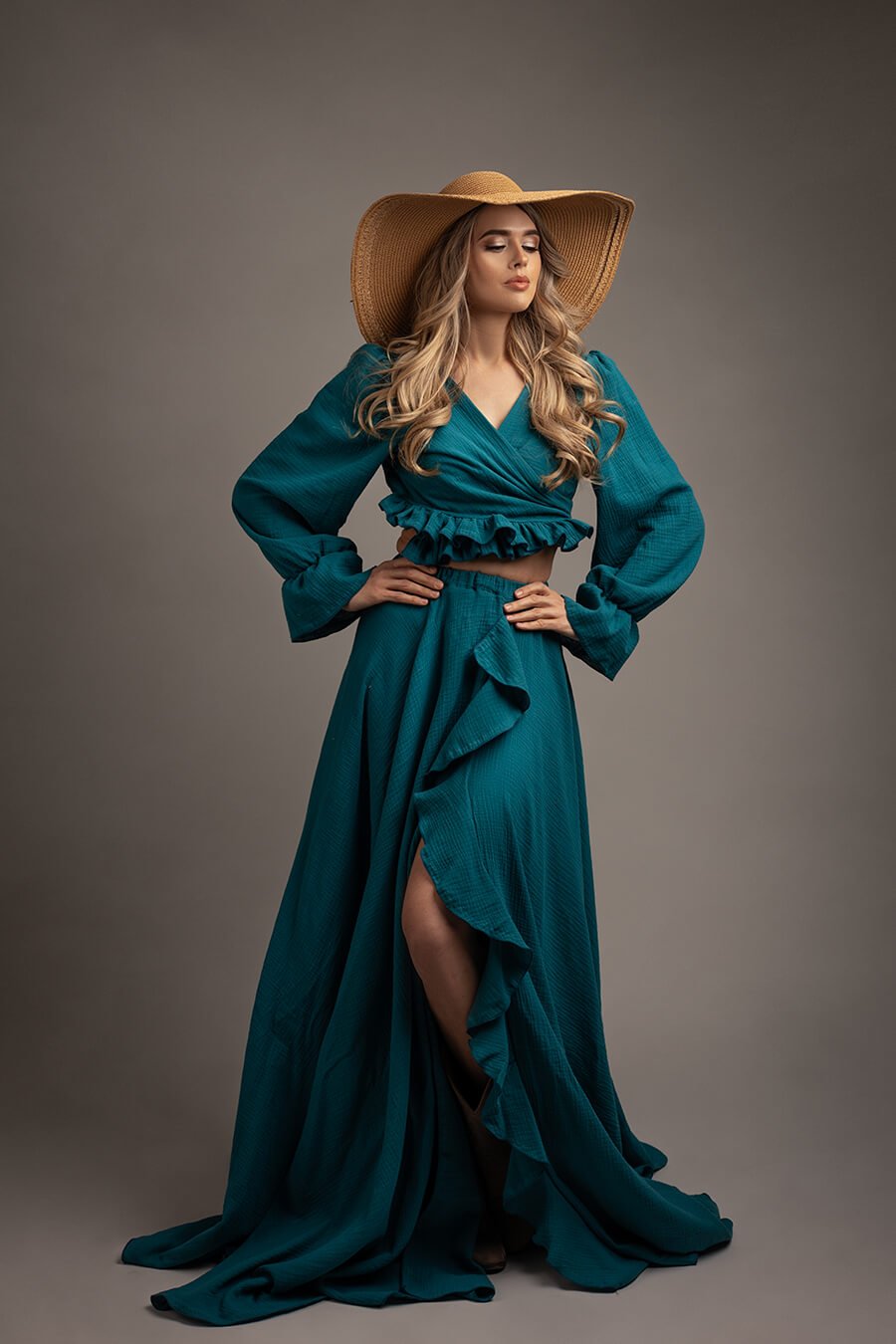 blond model poses in a studio wearing a boho western chic set in petrol color. she wears a top with ruffle details and long sleeves. she has a skirt with ruffle details to match the style and a brown hat on her head. 