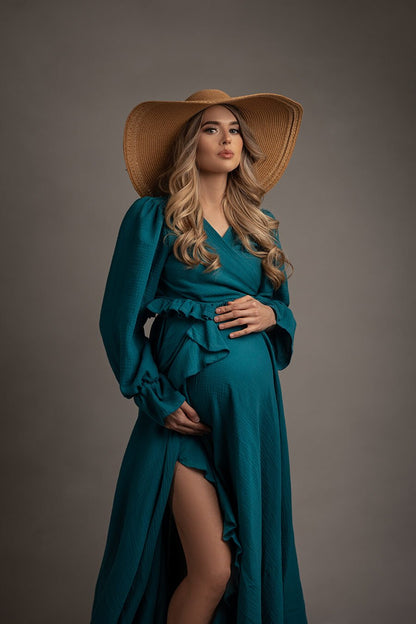pregnant model poses in a studio wearing a petrol boho western chic style dress with ruffle details and long sleeves. she has a brown hat to match the boho style.