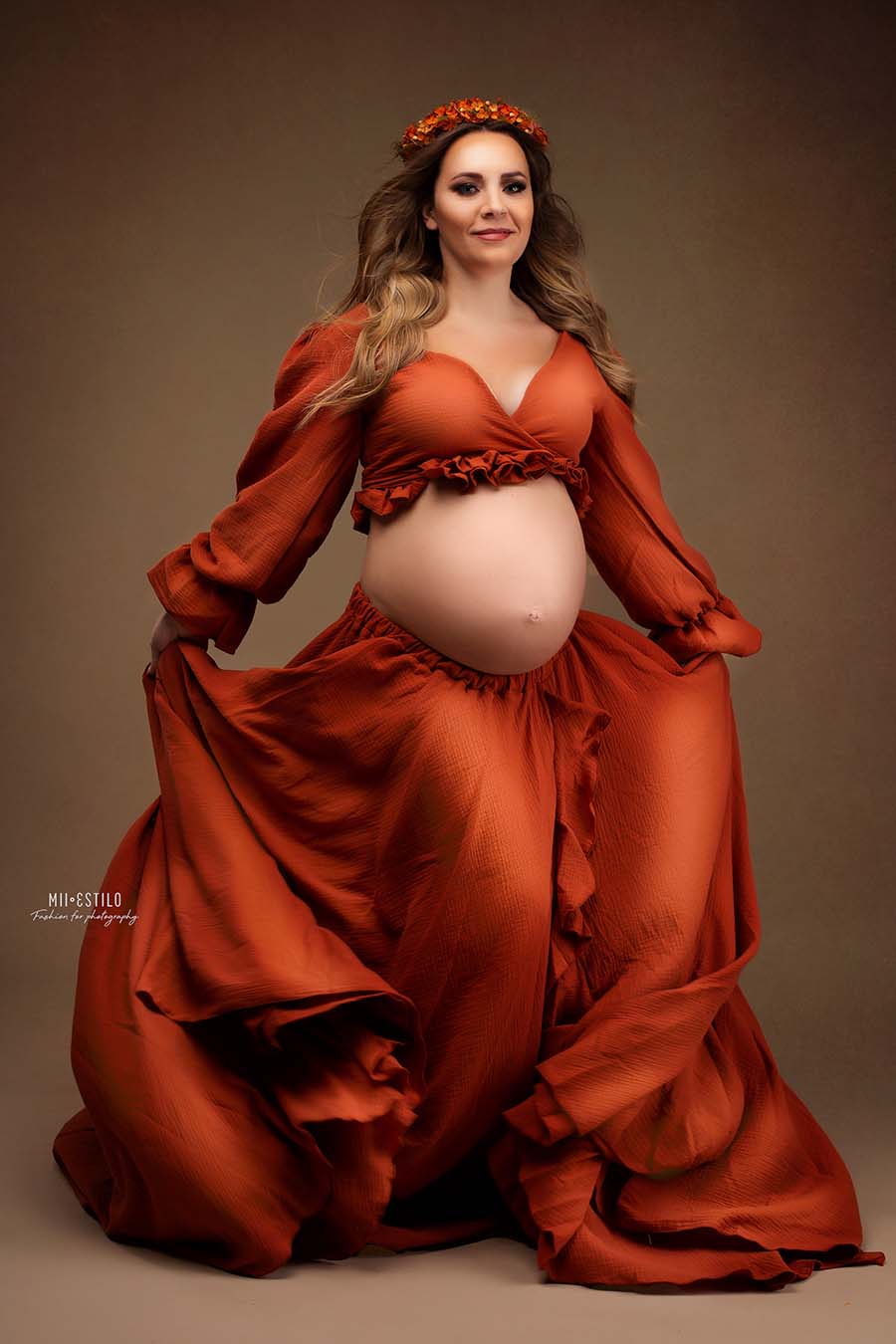 pregnant model poses in a studio wearing a rust orange set. She has a top with ruffle details, a low v cut neckline and bishop sleeves - and also a long circle skirt with ruffle details in the side split. she also wears an orange hear band to match the look. 