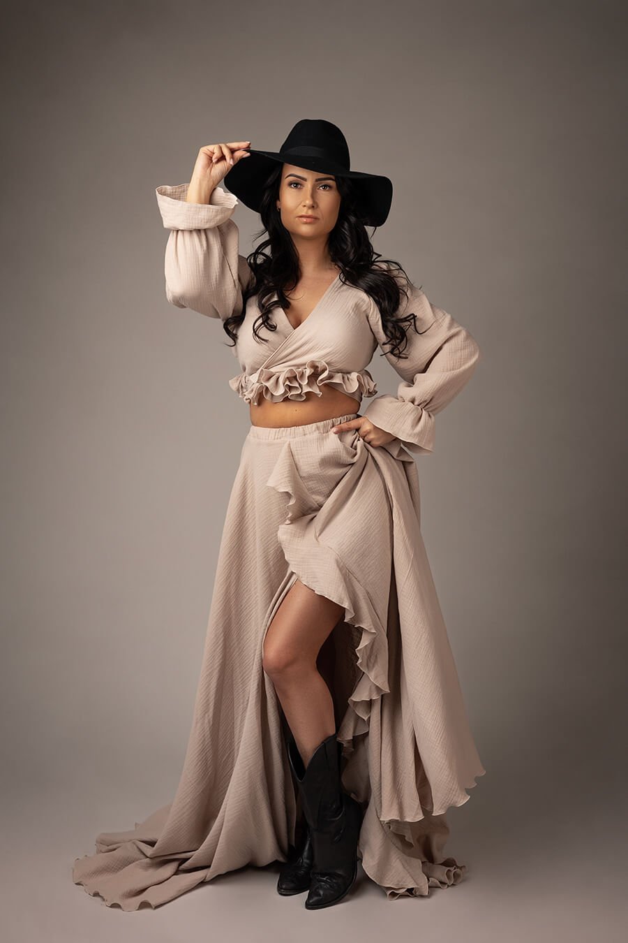 Brunette model poses in a studio wearing a sand set in boho chic style. she has a black hat and black boots to match the style. she is wearing a long skirt with ruffle details and a short top with long sleeves, also with ruffle details. 