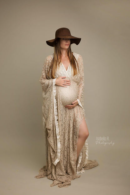 Brunette model poses in a studio during a maternity photo session. She wears a long dress made of lace and a matching hat. She looks to the camera while holding her bump with both hands. A bit of her leg can be seen through the split on the skirt.