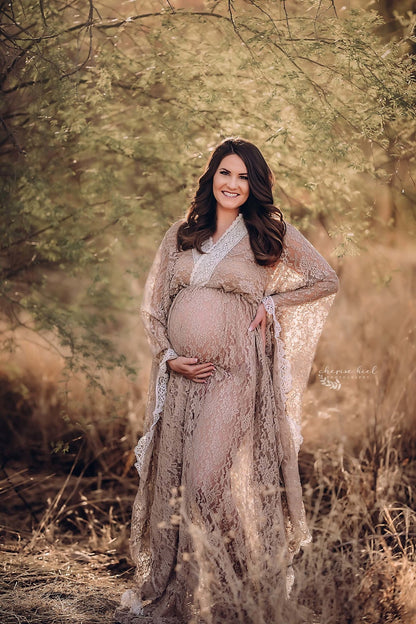 Brunette pregnant woman poses outside during a maternity photo session wearing a long dress made of lace from Mii-Estilo. She looks to the front and has one hand in her waist and the other holding her bump. The top is made of lace and features a low-v cut with kaftan sleeves. 