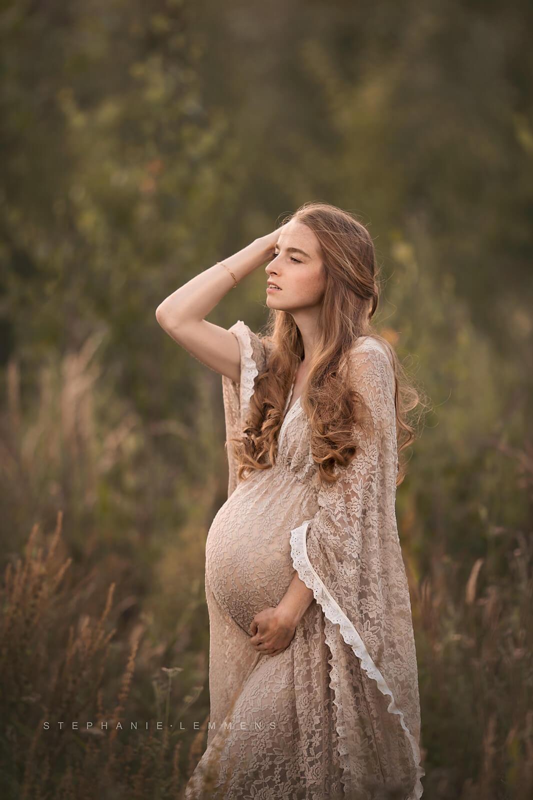 Blond pregnant model poses outside wearing a lace dress. The close up is made with her profile while she looks away, holding her bump with one hand and touching her hair with the other one. 