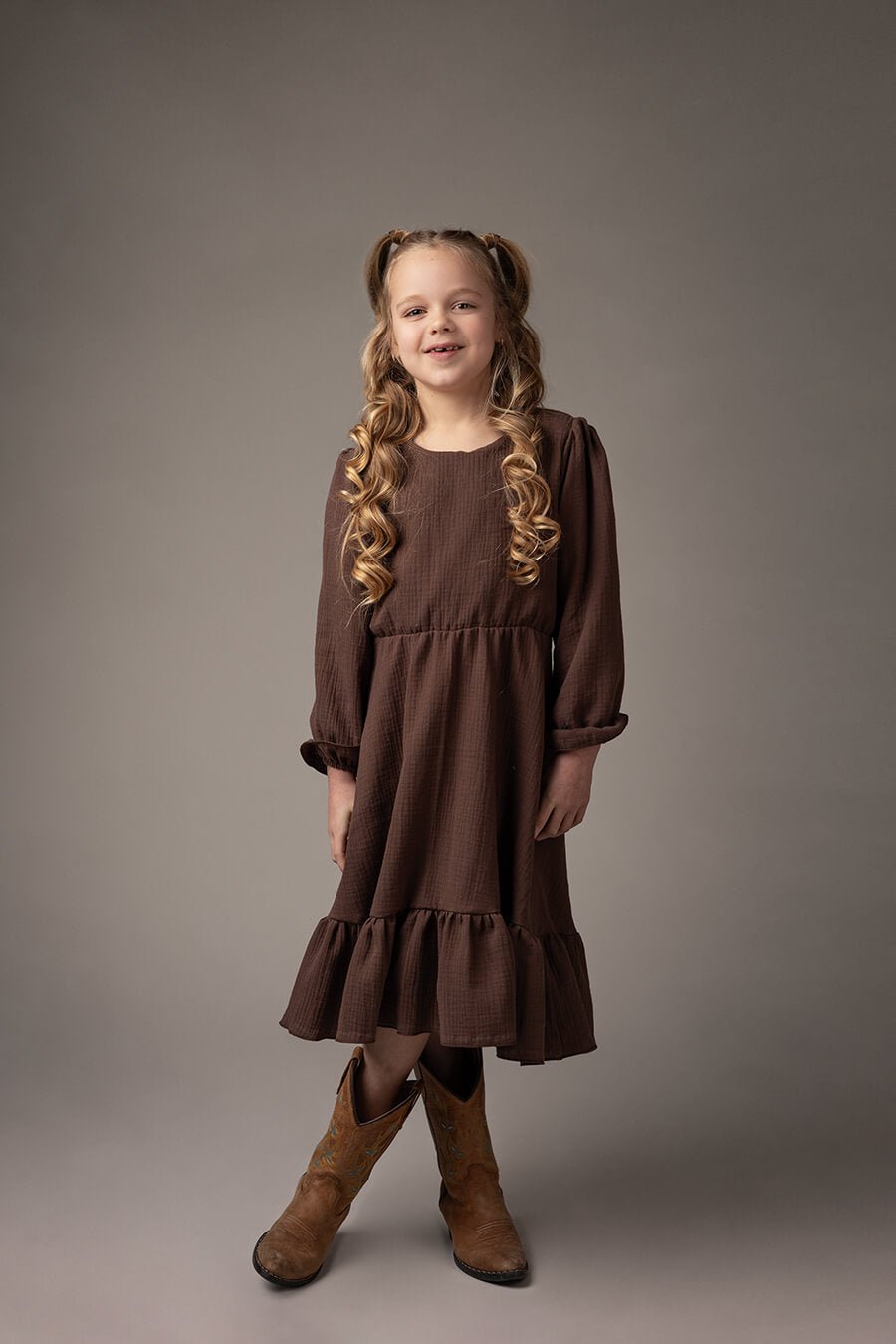 blond girl poses in a studio wearing a mid long chocolate dress in boho style. the dress has long sleeves and ruffle details. she has boots to match the western chic style.