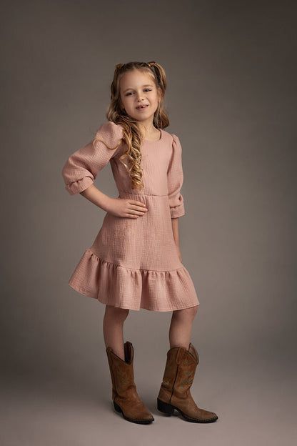 blond girl poses smiling to the camera during a boho western chic photoshoot in a studio. she has a dusty pink linen dress with ruffle details at the skirt and sleeves. she has brown boots to match the boho style. 