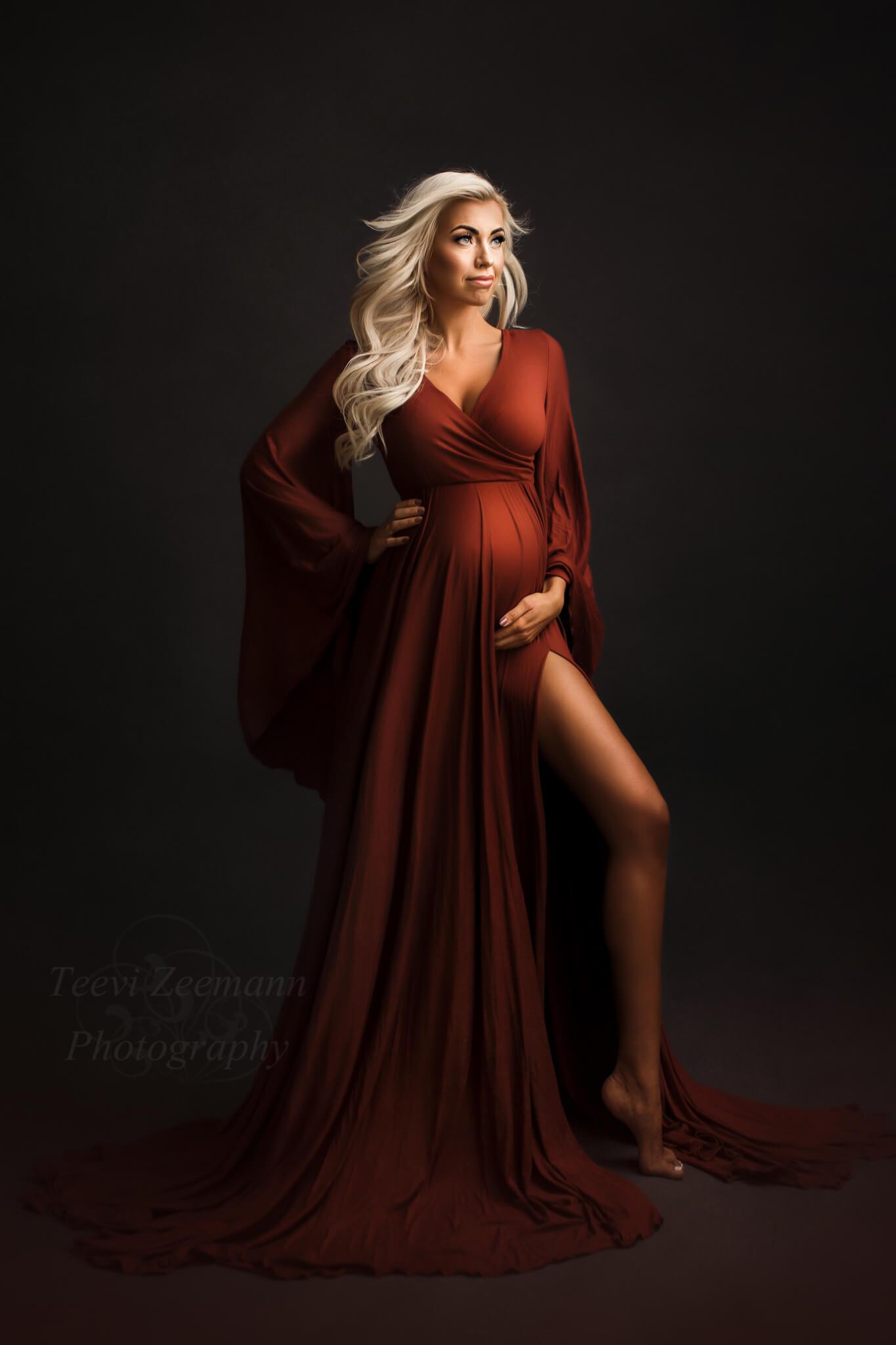 Blond pregnant woman poses in a studio during a maternity photoshoot. She is wearing a long maternity dress in Rust Orange color. The dress features a low v cut neckline with an adjustable sweetheart top and a long circle skirt with a split on it. The sleeves are long and in kaftan styke. 
