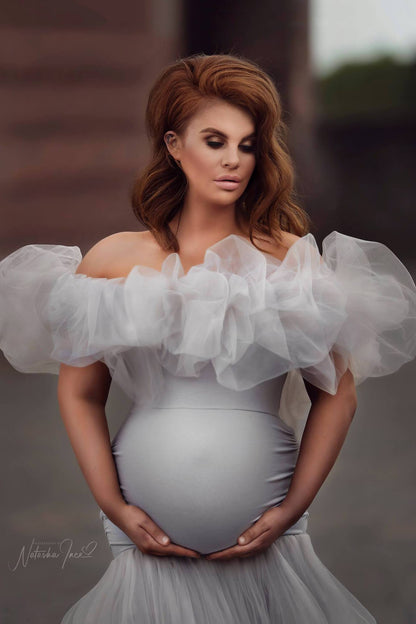 This pregnant model has red hair. She is looking down at the ground. the model is standing outside and has her hands underneath her belly. The dress has a off shoulder band made out of tulle fabric. The rest of the dress is tight. 