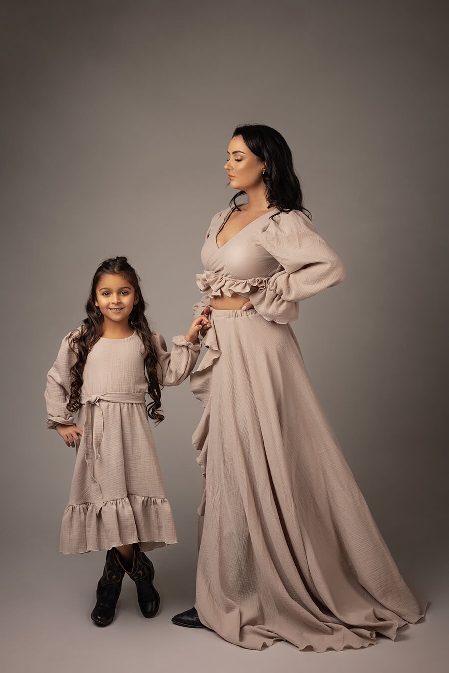 mom and daughter both with dark hair pose holding hands in a studio during a boho photoshoot. they are both wearing a sand outfit made of linen. both dresses have several ruffle details. the mom wears a long skirt and a top with long sleeves and an adjustable sweetheart top.