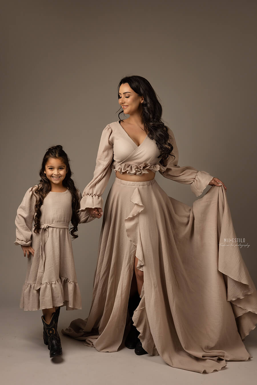 mom and daughter both with dark hair pose holding hands in a studio during a boho photoshoot. they are both wearing a sand outfit made of linen. both dresses have several ruffle details. the mom wears a long skirt and a top with long sleeves and an adjustable sweetheart top. 