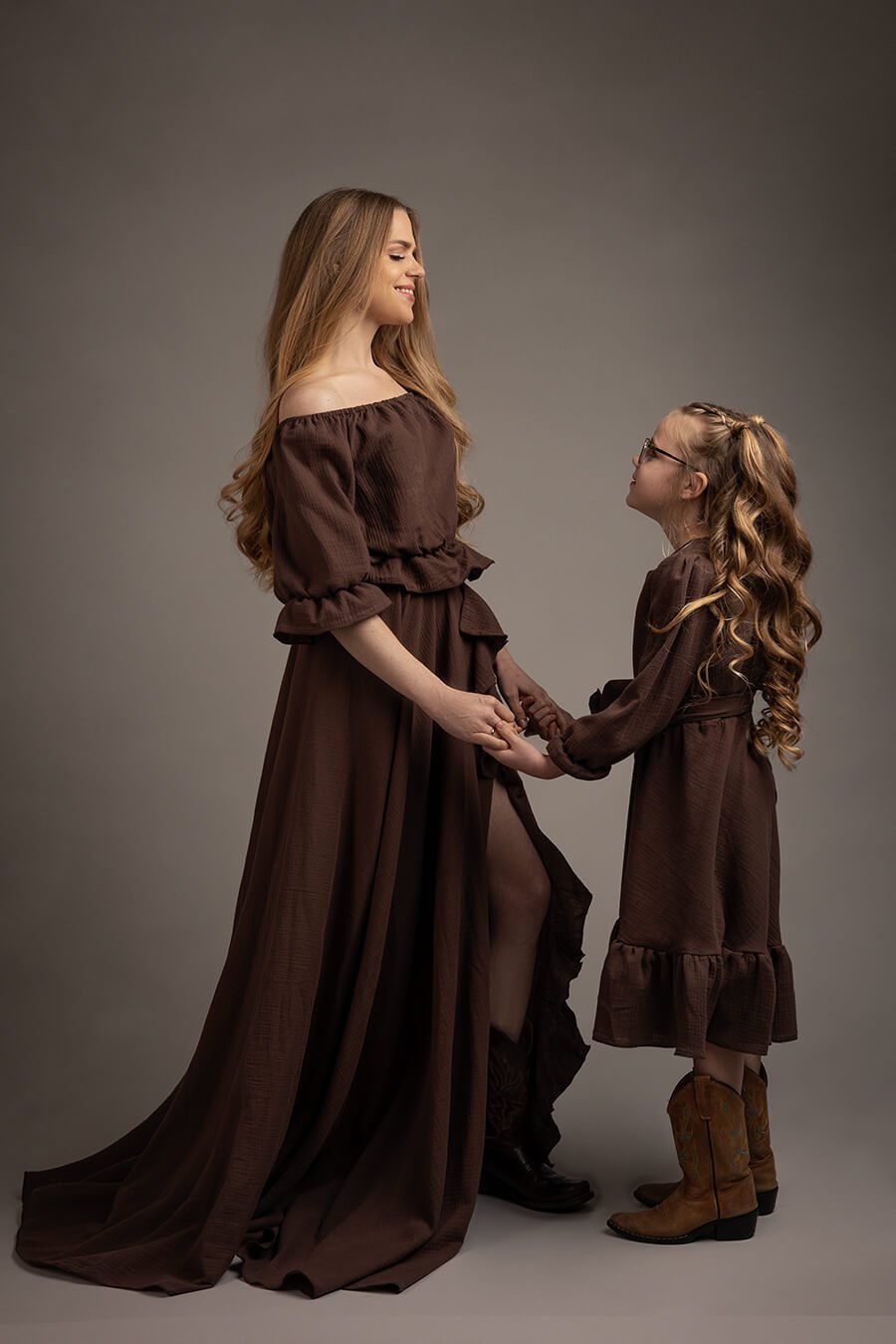 blond mom and kid are posing in a studio during a boho western chic photoshoot. they are both wearing chocolate color boho long dresses and boots to match the western style.