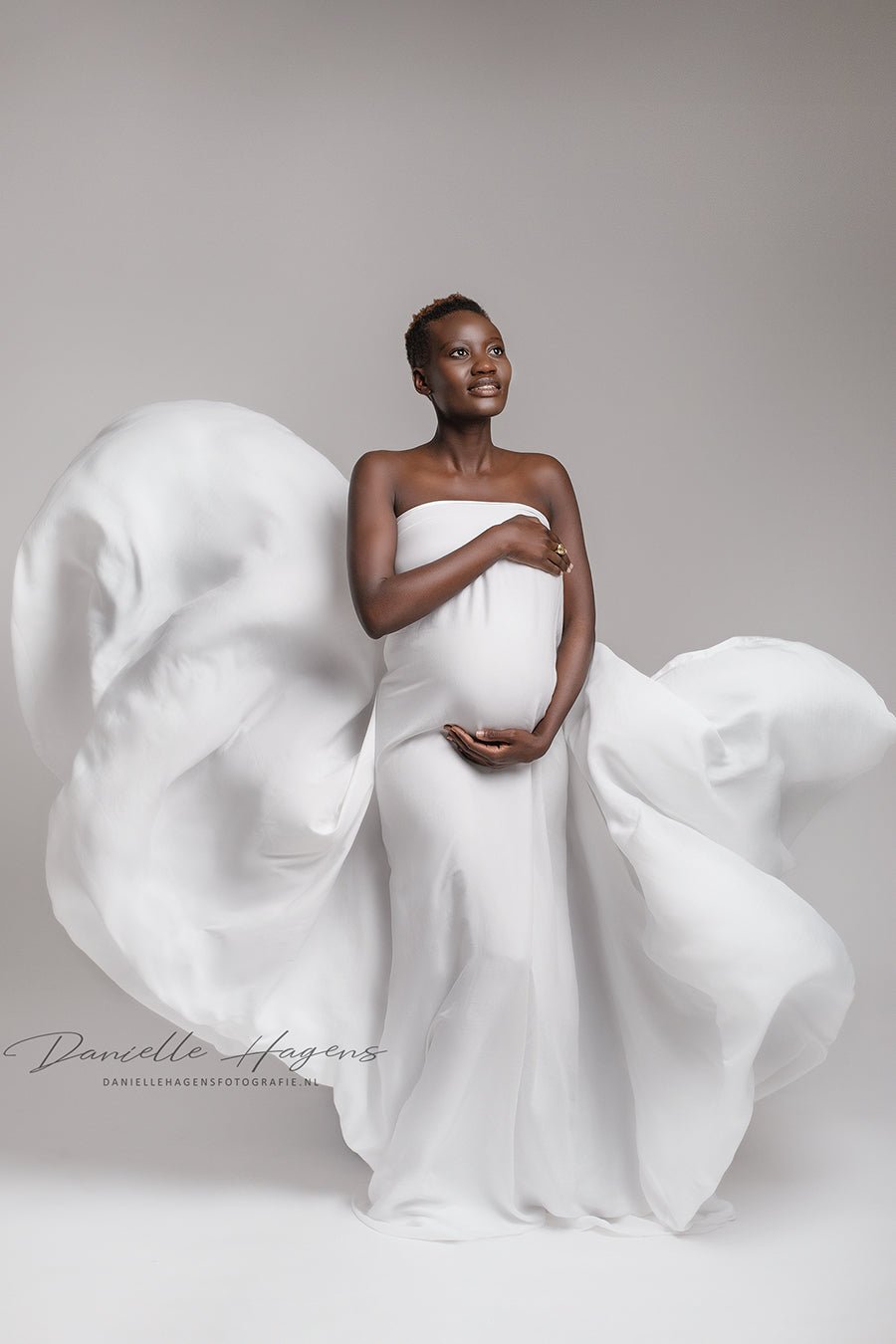 pregnant model poses covering her body with an off white silky scarf. she looks above white holds her belly bump.