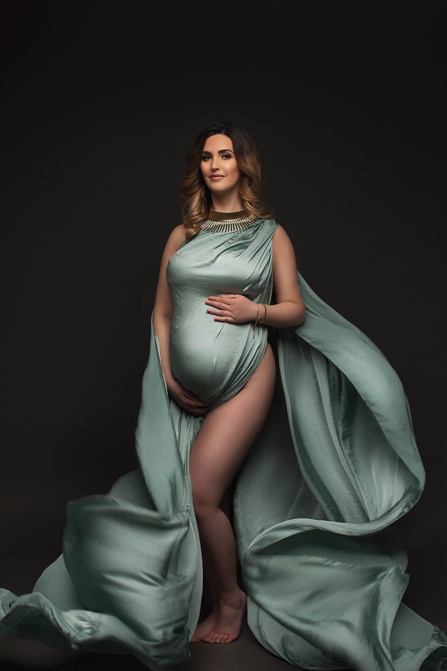 pregnant model poses wearing a draping fabric in azur color stylished as a dress. she has a big gold necklace to match the look.