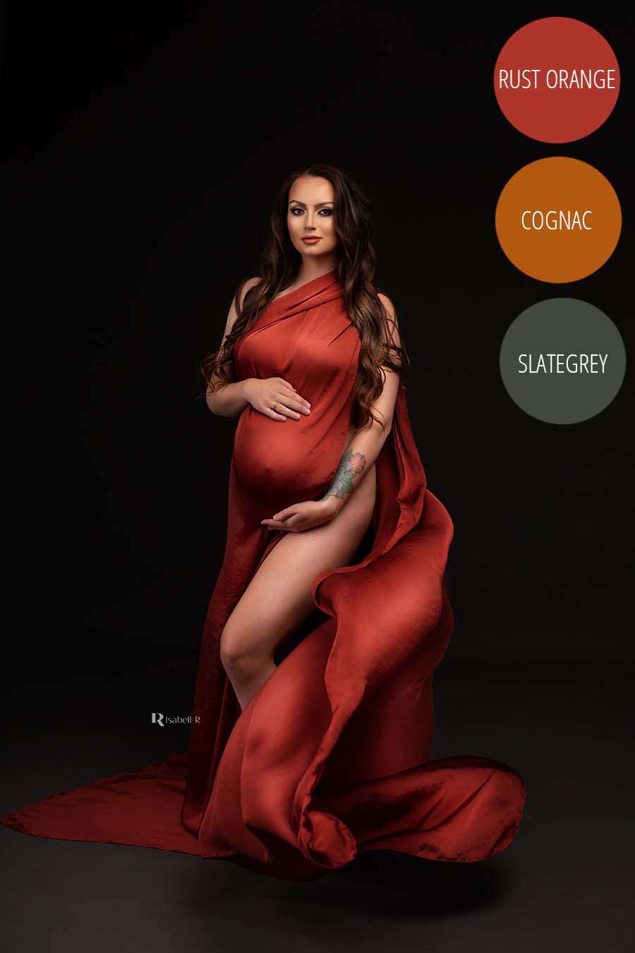 brunette model with long hair poses in a dark studio with a silk rust orange scarf wrapped around her body. she holds her bump with both hands. one of her legs can be seen.
