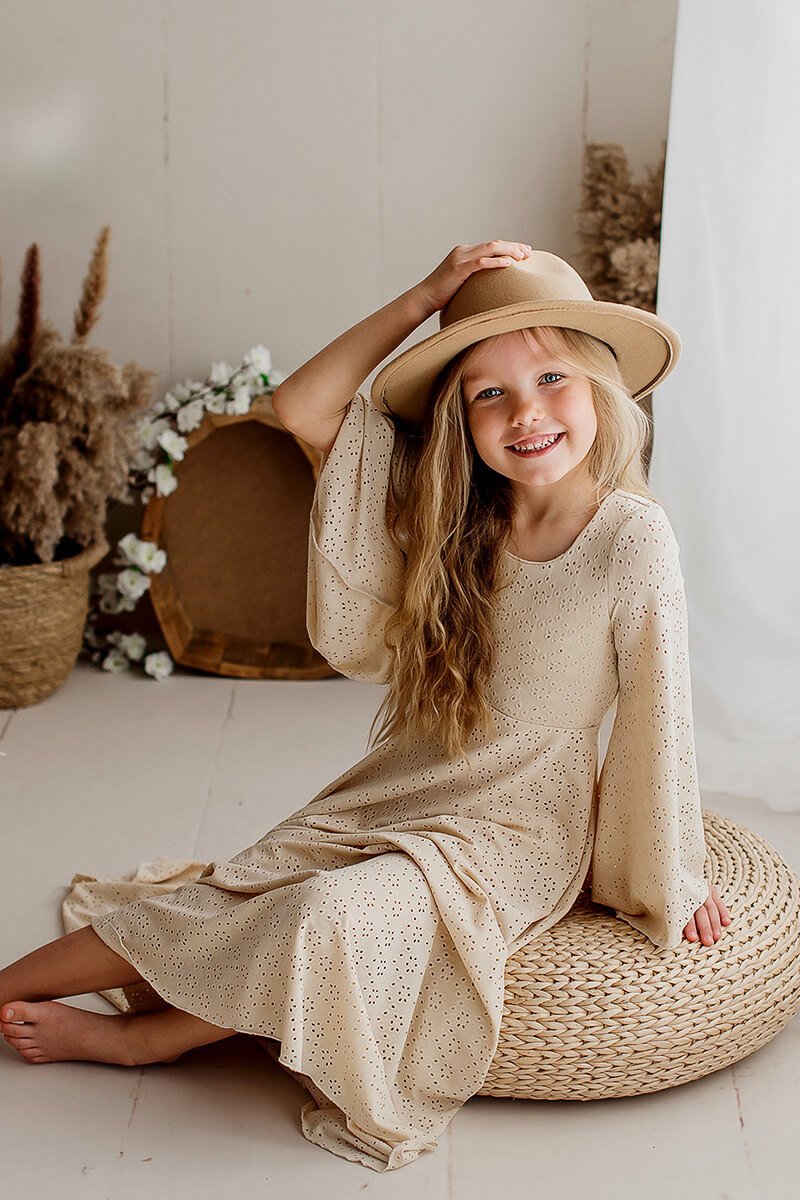 Little girl poses in a studio wearing a long dress made of brocante jersey in sand color. She stares and laugh to the camera and has a hat to match the style. 