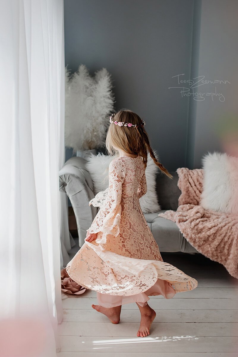 Little girl poses for a photoshoot wearing a lace dress in dusty pink and a flower crown. She is turning her back to the camera, in a way that the back from the dress can be seen.