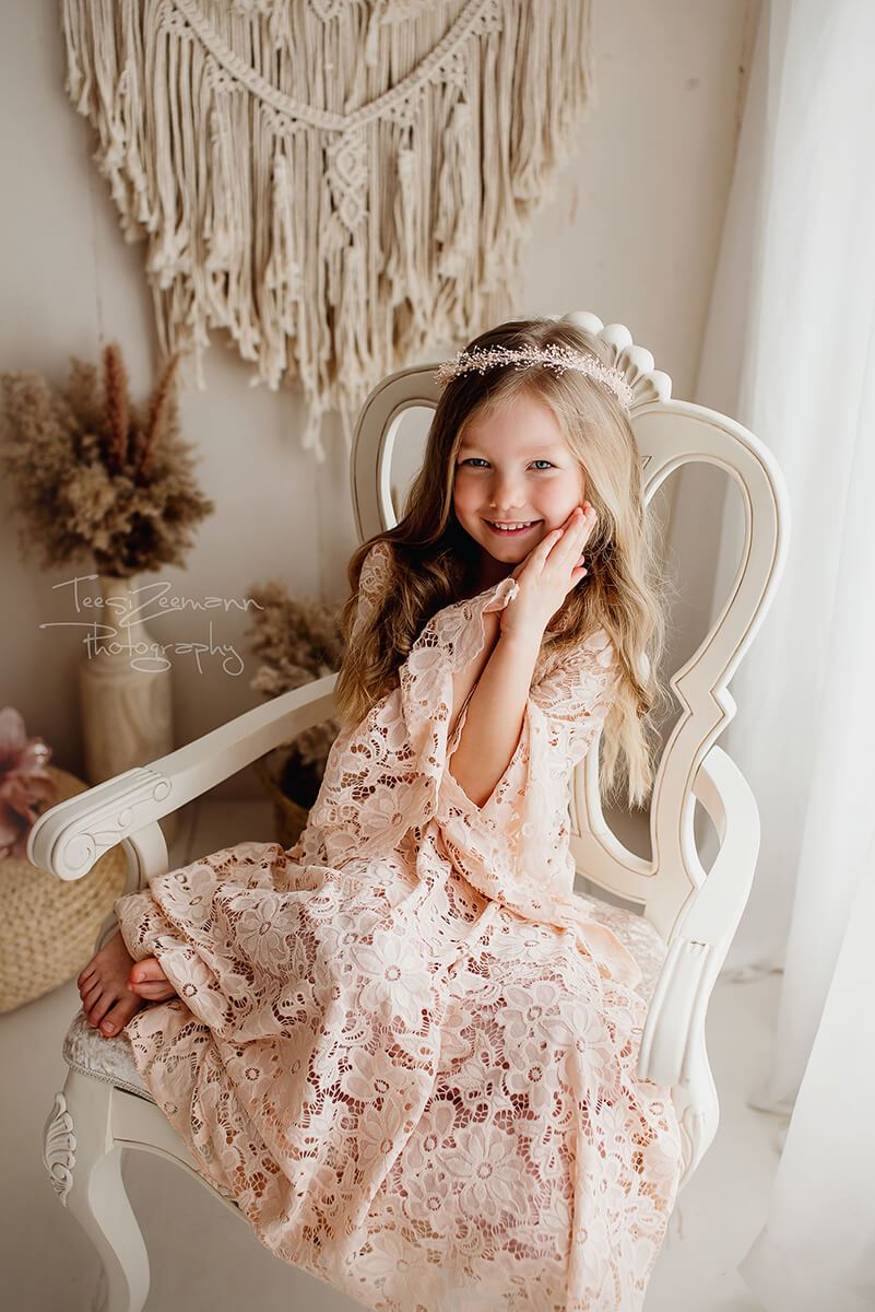Girls poses in a studio during a photo session. She wears a lace dress in dusty pink color and a flower crown. She is sitting on a white chair and has both of her hands on her cheek. She smiles and stares at the camera. 