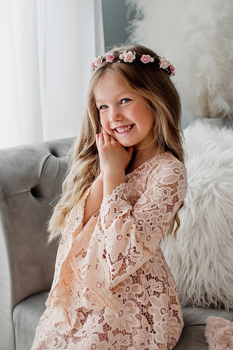 Girls poses in a studio during a photo session. She wears a lace dress in dusty pink color and a flower crown. She is sitting on a grey sofa and has both of her hands on her cheek. She smiles and looks away from the camera.