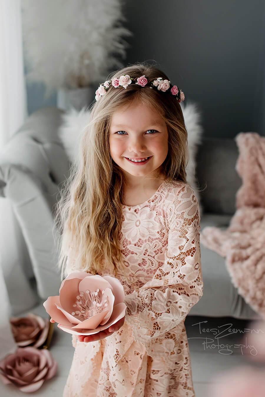 Little girl poses during a photo session holding a paper dusty pink flower and smiling at the camera. She is wearing a long lace dress in dusty pink and a matching flower crown. 