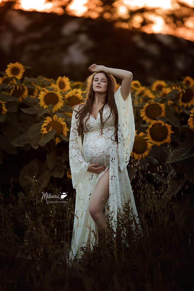 Pregnant model poses in a sunflower field. She has one hand on her bump and the other laying on the top of her head. Her eyes are closed and her head faces up. She wears an off white lace dress with a split where one of her legs can be seen. 