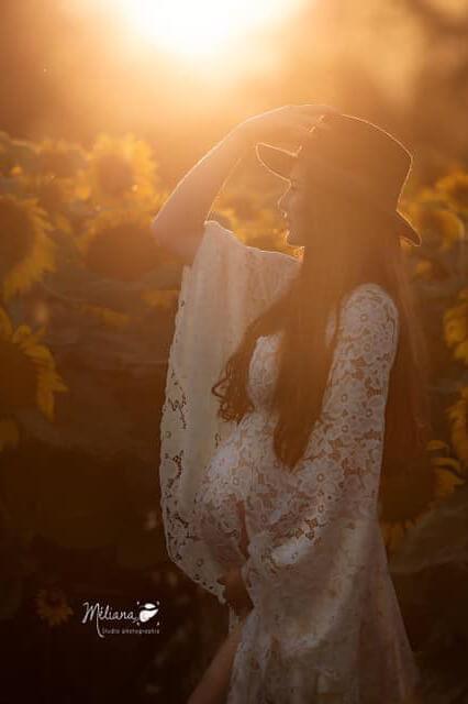 Pregnant model poses in a sunflower field. She has one hand on her bump and the other holding a hat. The sun makes gives the photo a sepia tone. She wears an off white lace dress with a split where one of her legs can be seen.