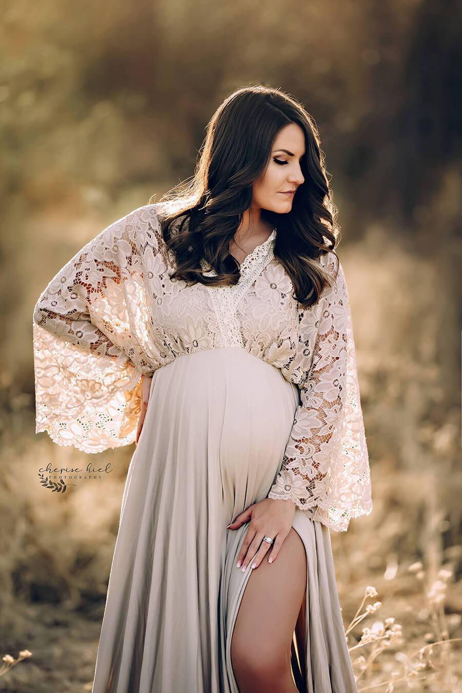 Brunette pregnant woman poses outside during a maternity photo session wearing a long dress made of jersey and lace from Mii-Estilo. She looks to the side and has one hand in her waist and the other on her legs. The top is made of lace and features a low-v cut with kaftan sleeves. One of her legs can be seen through the split form the skirt. 