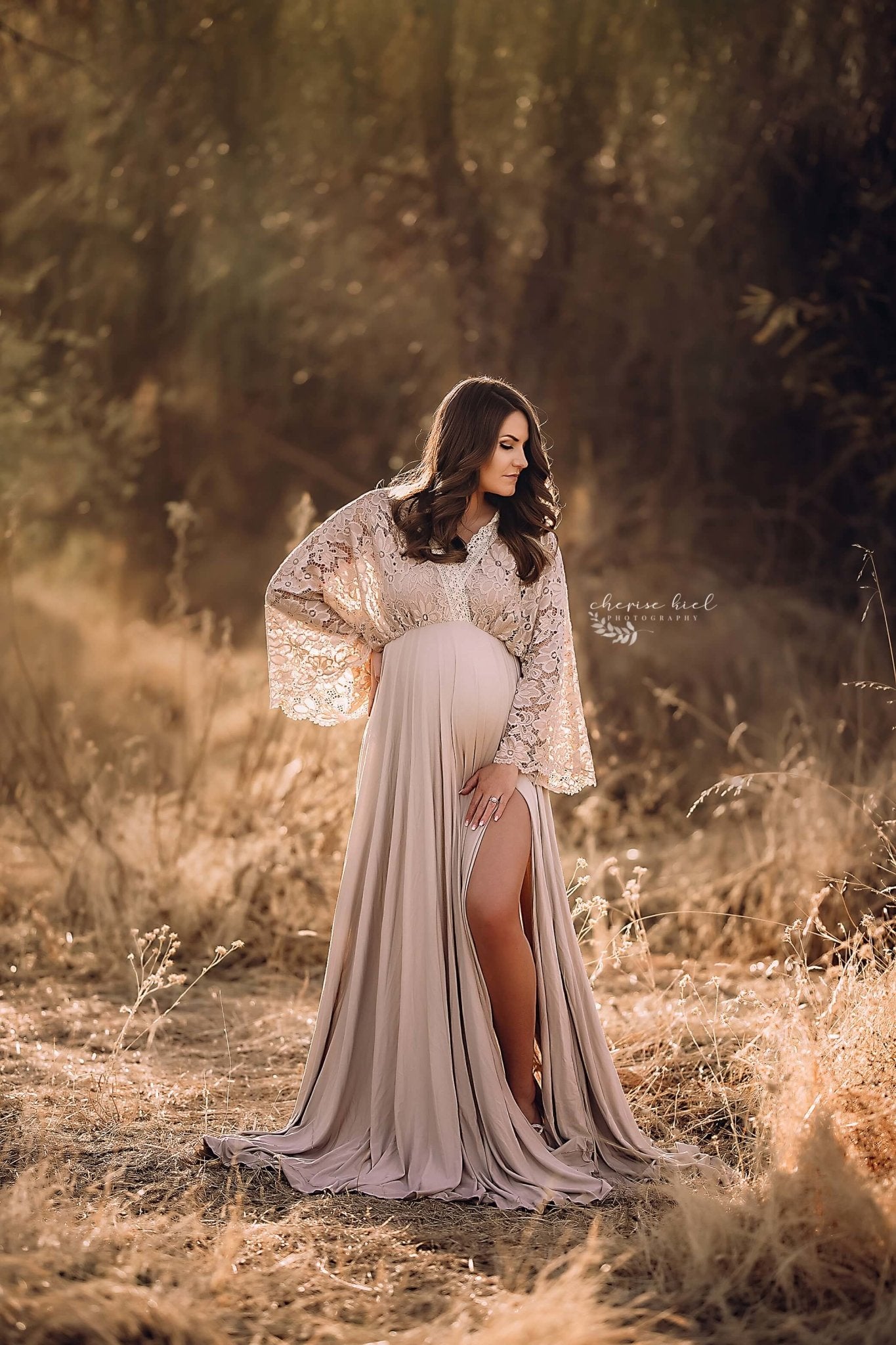Brunette pregnant woman poses outside during a maternity photo session wearing a long dress made of jersey and lace from Mii-Estilo. She looks to the side and has one hand in her waist and the other on her legs. The top is made of lace and features a low-v cut with kaftan sleeves. One of her legs can be seen through the split form the skirt. 