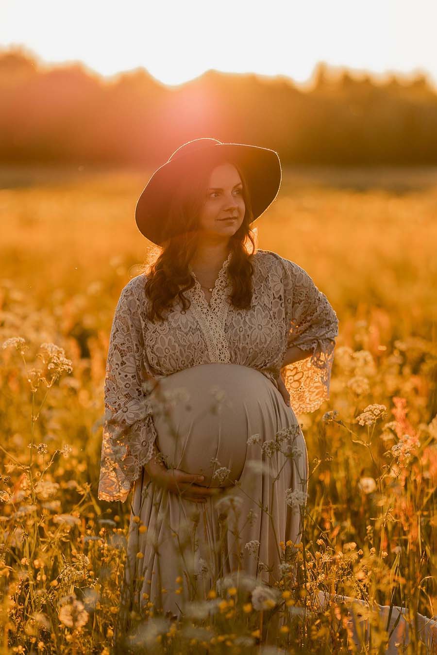 pregnant model poses outdoors during the golden hour wearing a boho style dress made of lace and jersey in sand color. she has a hat to match the western chic style.