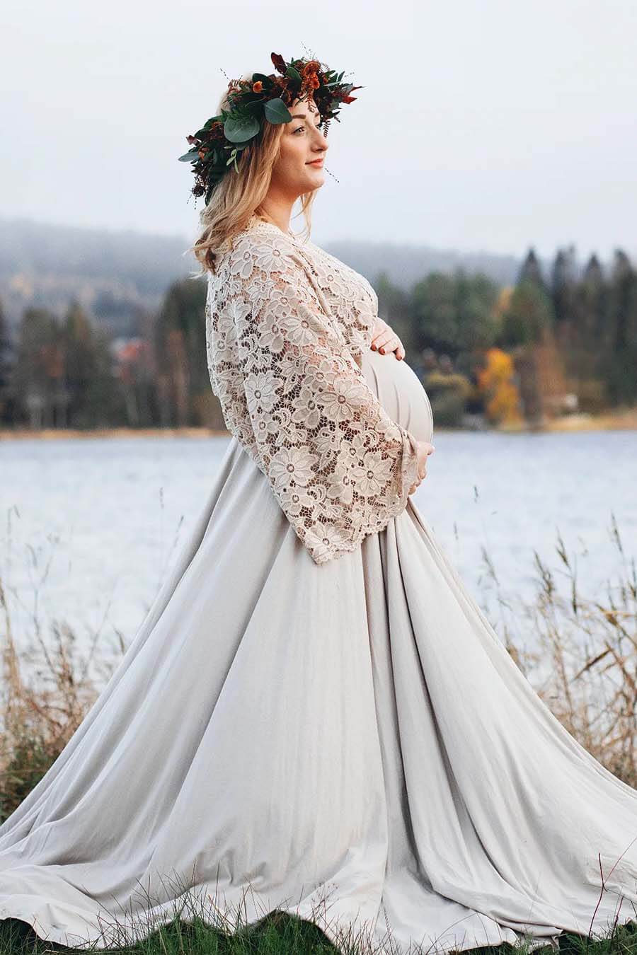 pregnant model poses outside nearby the water. she is wearing a maternity dress in sand color - the dress is made in lace and jersey. the top has long kaftan style sleeves and is made totally in lace while the long circle skirt is made in jersey. the model has a flower crown.