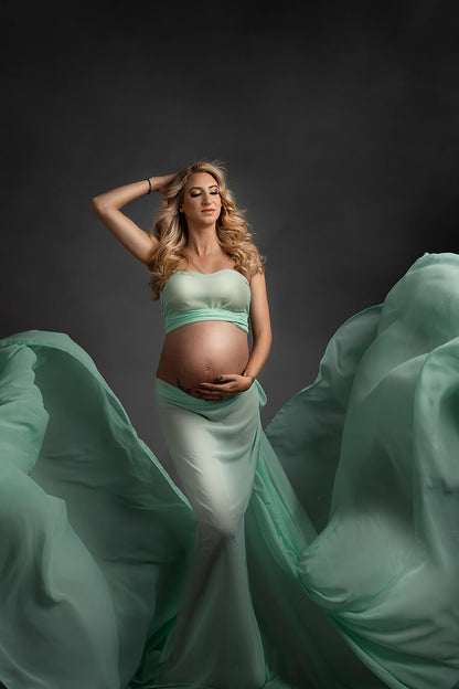 blond pregnant model poses in a studio wearing a mint green draping fabric wrapped around her body. Her belly in uncovered.