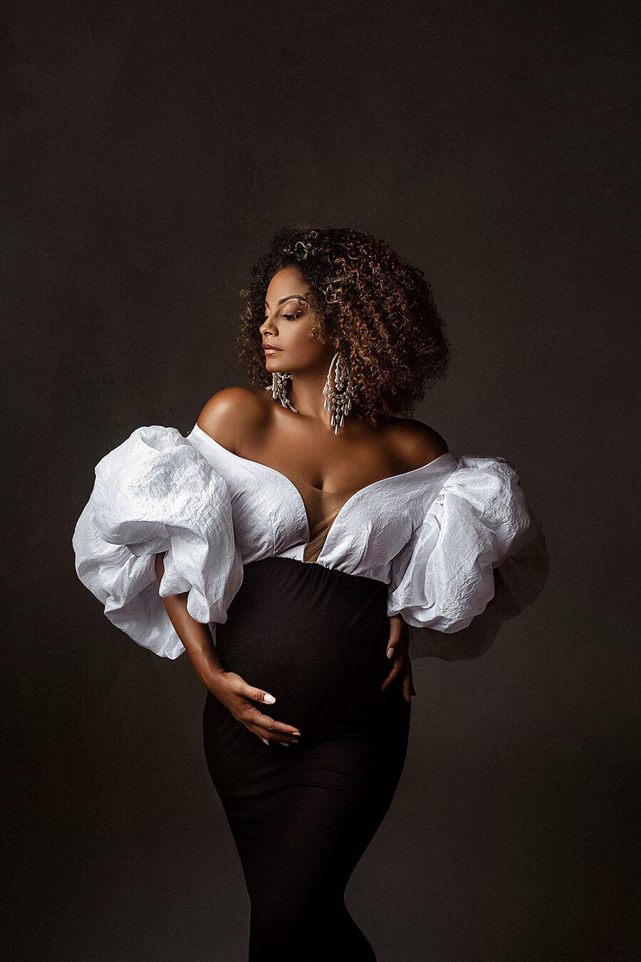 black pregnant model poses in a studio wearing a red carpet dress in 2 colors. the top is made of taft with exaggerated puff sleeves and the mermaid style skirt is black. 