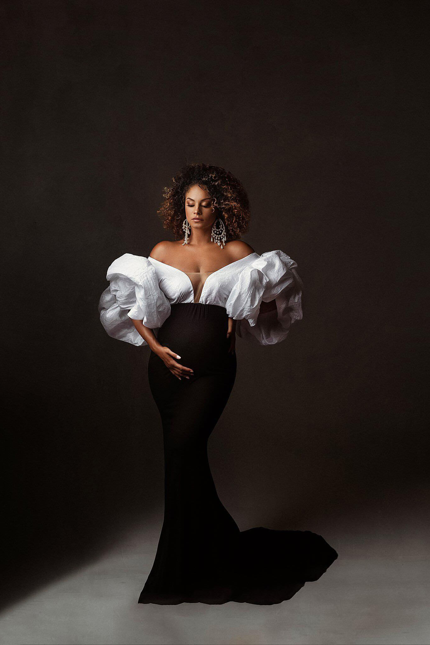 black pregnant model poses in a studio wearing a red carpet dress in 2 colors. the top is made of taft with exaggerated puff sleeves and the mermaid style skirt is black. 