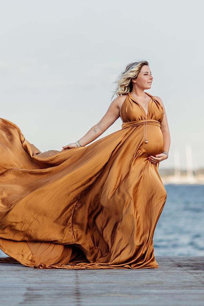 blond pregnant model poses outside by the water as a background. she is wearing a camel silky dress with a long skirt. 