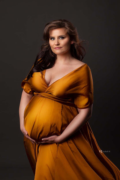 A pregnant woman is wearing a long dress. The dress is in the color cognac. The dress has a lot of fabric and is shiny. The top has a v-neck and short sleeves.