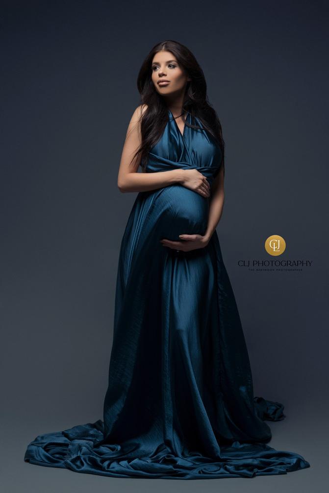A pregnant woman is wearing a long petrol dress. The dress has no sleeves. The woman is holding her belly