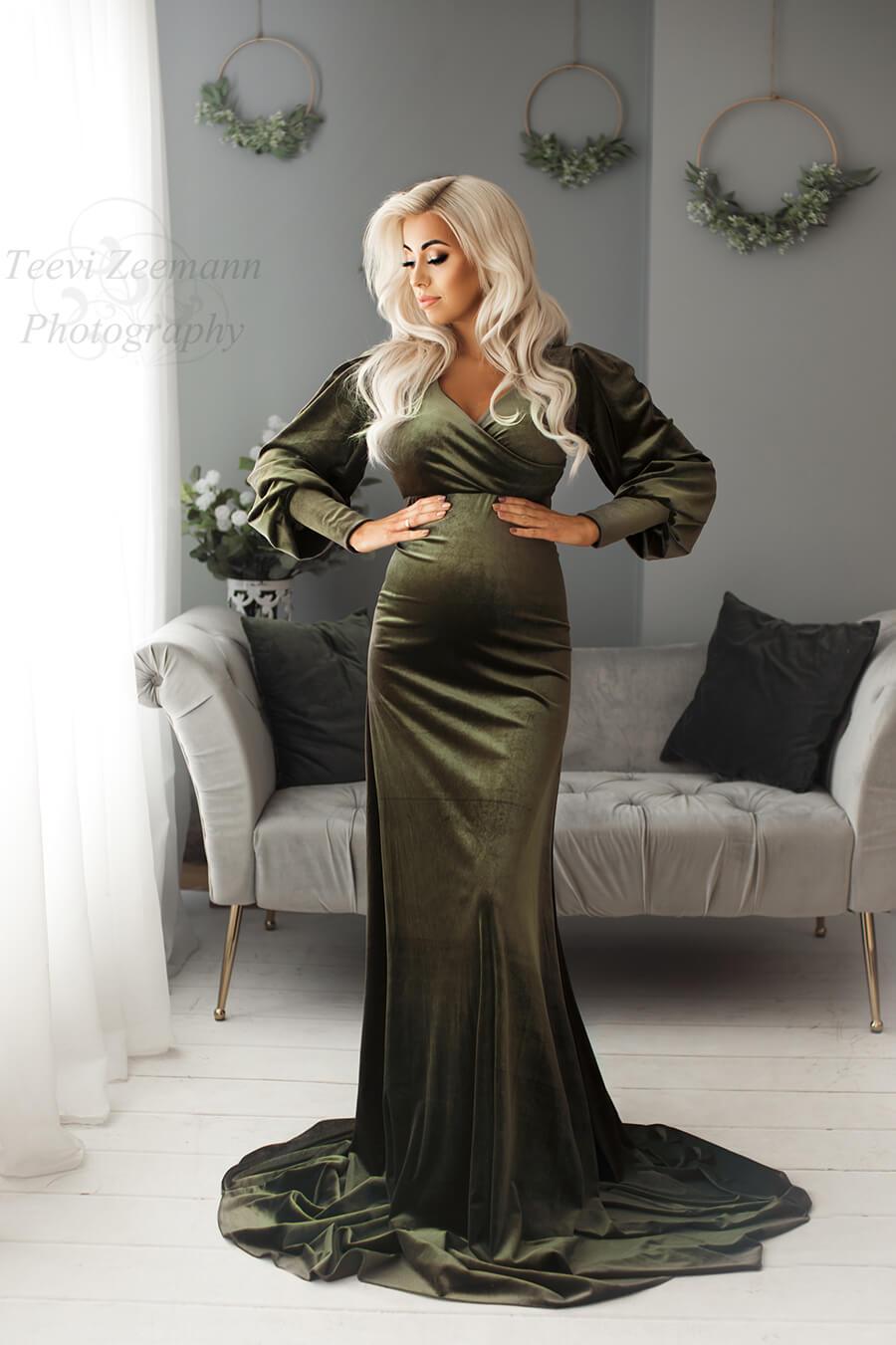 The woman is standing in a living room. She is wearing a long olive green dress with a sweetheart top. The dress is velour and the woman has long blonde hair and she is pregnant