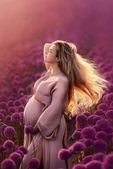 Pregnant model poses outside in a field with flowers. She is wearing a silky long dress in dusty pink color. The model touches her long blond hair and has her eyes closed. with the other hands she touches her bump. 