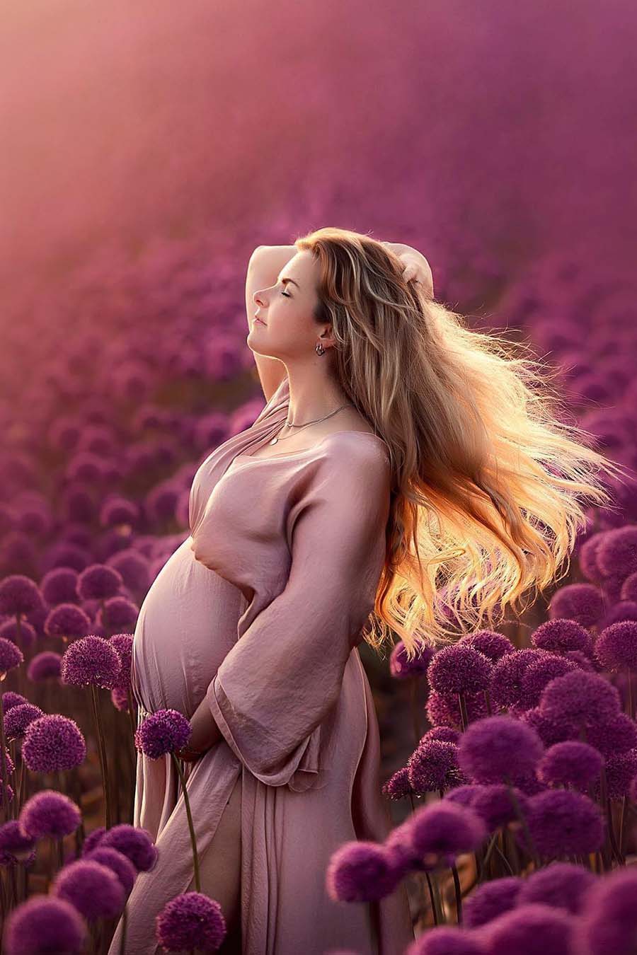 Pregnant model poses outside in a field with flowers. She is wearing a silky long dress in dusty pink color. The model touches her long blond hair and has her eyes closed. with the other hands she touches her bump. 