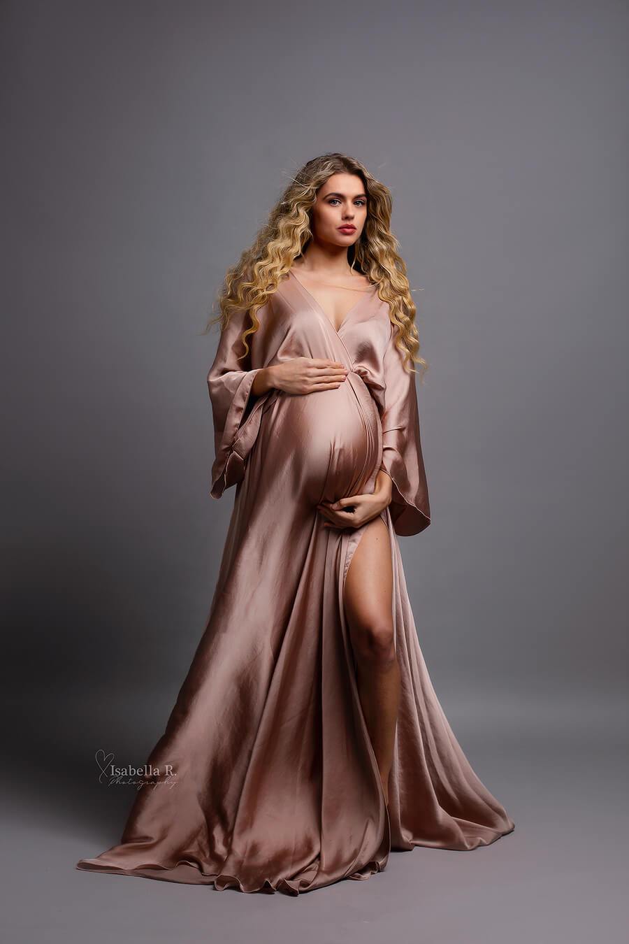 pregnant model with long wave hair poses in a studio with a long silky dress in dusty pink color. she is staring to the camera and touching her bump with both hands. one of her legs can be seen through the split in the skirt. 