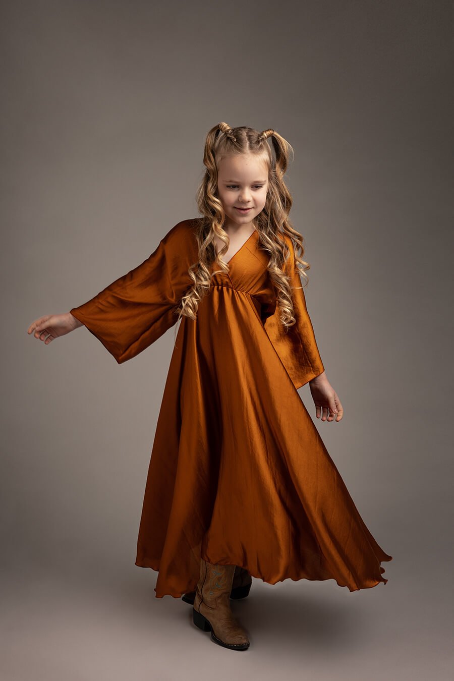 blond girl poses in a studio during a boho chic photoshoot. she is wearing a long silky cognac dress in a boho chic style and wears boots to match the look. 