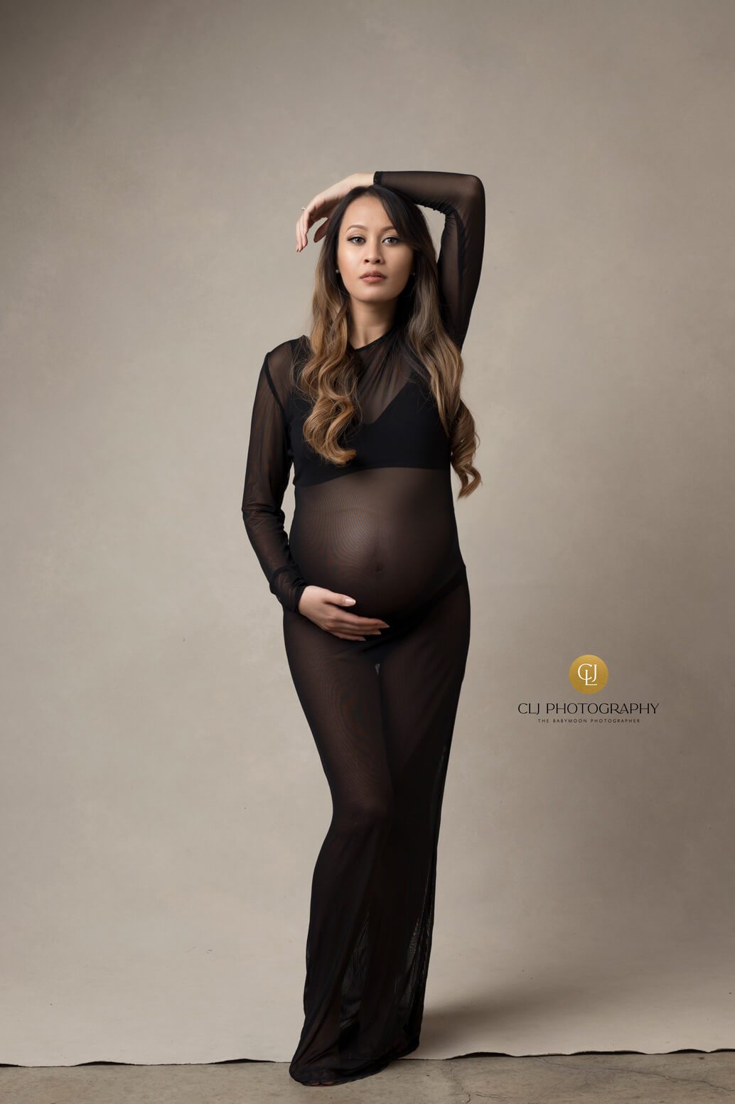 Model poses in a studio during a maternity photoshoot. She wears a transparent tight black dress and faces forward. One of her hands lays on her head and the other holds her bump. 