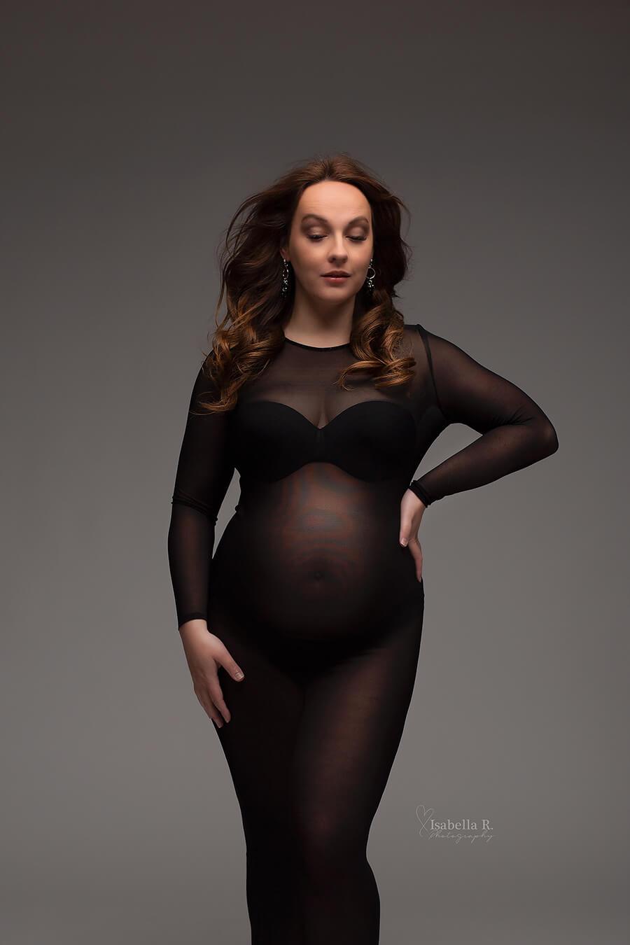 Model poses in a studio during a maternity photoshoot. She wears a transparent tight black dress and faces forward but has her eyes closed and her hands are laying on her waist and leg. 