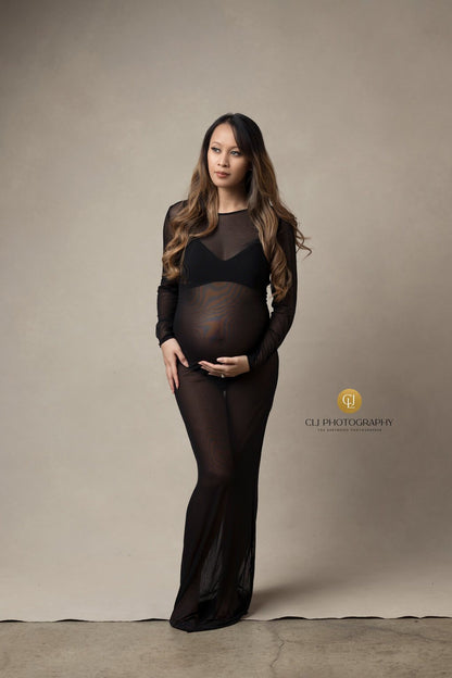 Model poses in a studio during a maternity photoshoot. She wears a transparent tight black dress and faces forward whole holding her bump with one of her hands.