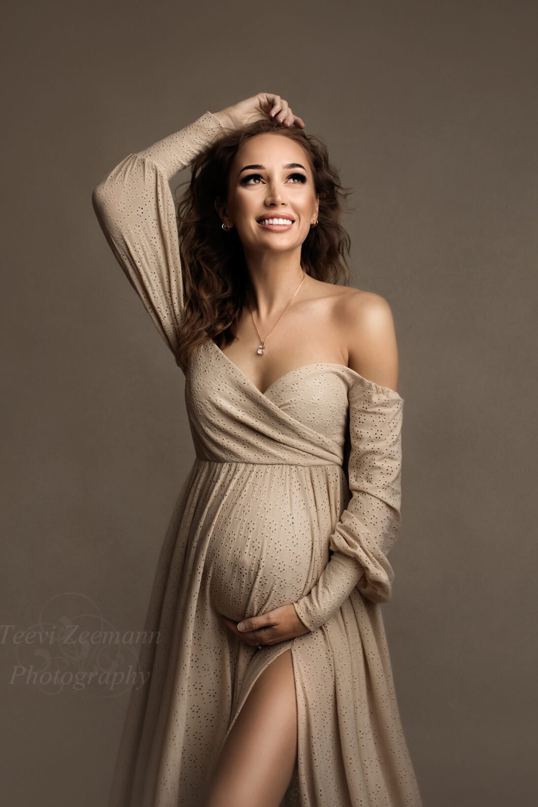 Brunette pregnant model poses for a studio maternity photo shoot. She wears a Mii-Estilo bohemian styled dress: the Crocus Dress in Sand color. One of her hands is above her head while the other one holds her bump. The sweetheart top is adjustable and the dress has long sleeves and off the shoulder. The skirt has a split - the dress is made of brocante lace. The dress can be partially seen in the photo and the model faces the camera with her eyes open.