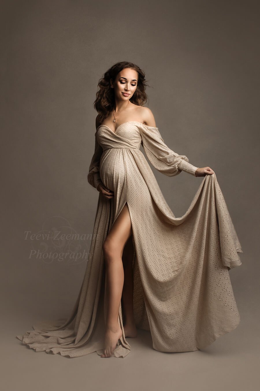 Brunette pregnant model poses in a studio for a maternity photo session. She wears a Mii-Estilo Crocus Dress in Sand that can be completely seen in the picture. The dress is made of brocante lace and features off the shoulder and adjustable sweetheart neckline. The model has her eyes closed and faces the side. She holds the skirt from the dress with one hand and her bump with the other.
