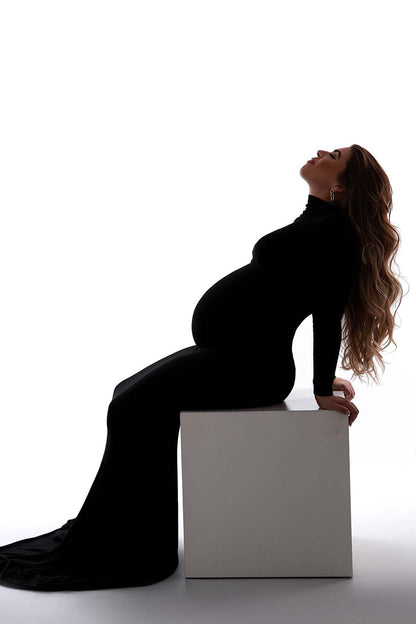 A pregnant woman with long brown hair is sitting on a white block in the photostudio. She is leaning on her back hands and is wearing a long black dress. The dress has a tight fit . It has a turtle neck and long sleeves.