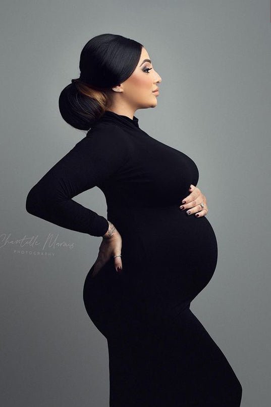 Pregnant model poses on her side in a studio during a maternity photo session. She holds her back and bump. She wears a long and tight dress featuring turtle neck and long sleeves. Her eyes are open and she looks away.