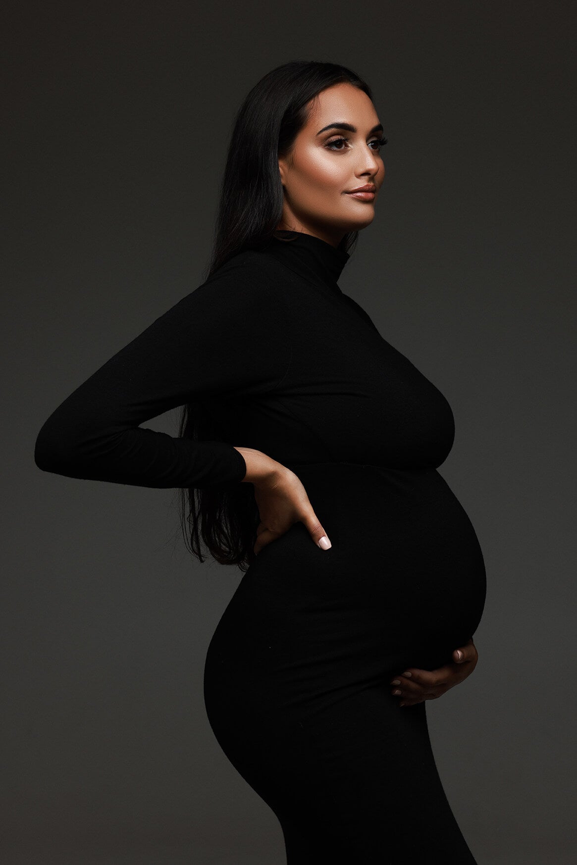 portrait photo of a pregnant model posing in a studio wearing a long tight black dress with a turtleneck neckline and long sleeves.